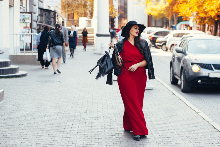 Young stylish woman wearing red maxi dress, black leather jacket and hat walking on the city street in autumn. Fall fashion, elegant look. Plus size model.